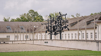 Auf einem kargen Platz steht vor einer Häuserreihe eine moderne Skulptur. Darin bilden menschliche Körper einen großen Ball.