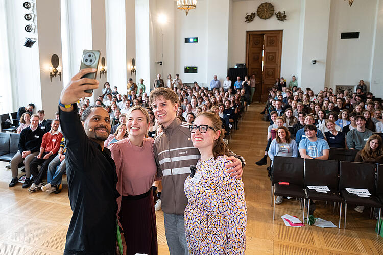 Vier Menschen machen auf einer Bühne ein Selfie mit dem Publikum im Hintergrund