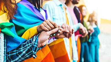 Zwei Frauen mit unterschiedlichen Hautfarben halten sich die Hände. Sie tragen queere Regenbogenflaggen um die Schultern.