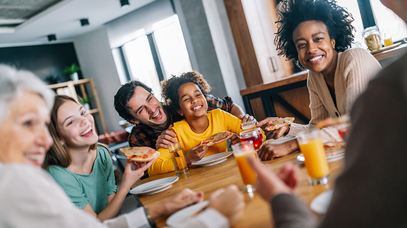 Menschen unterschiedlichen Alters und Hautfarbe sitzen gemeinsam lachend an einem Tisch und essen Pizza. 