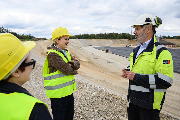 Katharina Schulze und Dr. Andreas Bauer im Gespräch, im Hintergrund das ausgehobene, aber noch leere Oberbecken des Pumpspeicherkraftwerks. Beide tragen neongebe Warnwesten und Bauhelme.