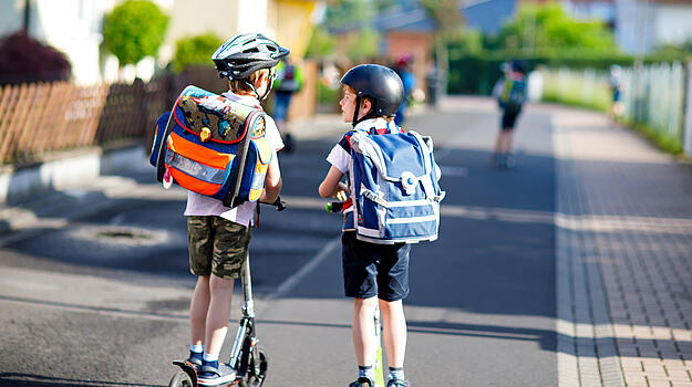 Zwei Schulkinder mit Helm und Schulranzen fahren mit ihren Rollern eine Straße entlang.