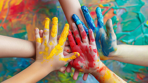Mit bunter Farbe bemalte Kinderhände stapeln sich vor einer bunten Wand übereinander.
