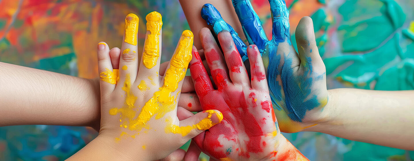 Mit bunter Farbe bemalte Kinderhände stapeln sich vor einer bunten Wand übereinander.
