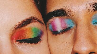 Das Bild zeigt einen Ausschnitt von den Gesichtern zweier Menschen, die eng aneinander liegen. Die Linke trägt Regenbogen-Lidschatten. Der Lidschatten der rechten Person hat die Farben blau, rosa, weiß, rosa, blau. 