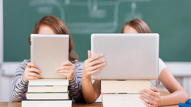 Zwei Kinder hinter Laptop und Tablet vor einer Tafel im Klassenzimmer.