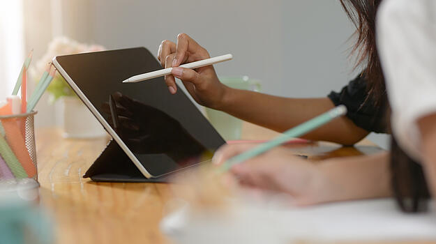 Hand tippt mit digitalem Stift auf Tablet. Daneben steht ein Stiftehalter mit Stiften.