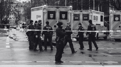 Das schwarz-weiß Bild zeigt eine Gruppe Polizisten steht vor Krankenwägen hinter einem Absperrband.