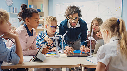 Junger Mann erklärt Kindern etwas an einem Tablet. Kinder sitzen im Kreis um den Tisch herum. Auf dem Tisch stehen kleine Windräder.