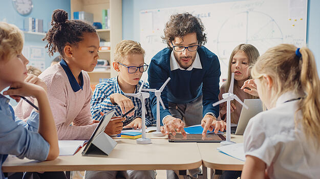 Junger Mann erklärt Kindern etwas an einem Tablet. Kinder sitzen im Kreis um den Tisch herum. Auf dem Tisch stehen kleine Windräder.