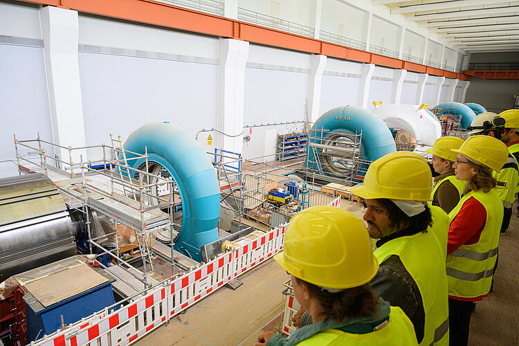 Eine Gruppe Abgeordneter, gekleidet in neongelbe Warnwesten und Bauhelme, blickt von einem Balkomn in die Maschinenhalle des Pumpspeicherkraftwerks. Zu sehen sind vier große Turbinen, umgeben von Baugerüsten und Absperrungen.
