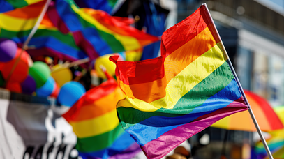 Im Vordergrund weht eine Regenbogenflagge. Verschwommen im Hintergrund wehen weitere Flaggen und bunte Luftballons.