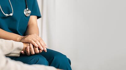 Person in dunkelblauer Uniform und Stethoskop um den Hals hält Hand von einer Person mit beige-farbigen Oberteil