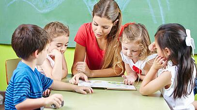 Junge Frau sitzt mit 4 Kindern am Tisch und liest ihnen vor. Im Hintergrund ist eine Tafel.