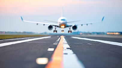 Scharf und im Vorderrund ist eine Landebahn aus Asphalt zu sehen. Unscharf setzt im Hintergrund ein Flugzeug auf die Landebahn auf.