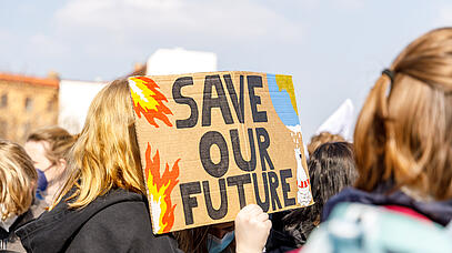 Einige junge Menschen sind von hinten zu sehen. Eine Hand hält ein Pappschild mit den Worten "Save our Future" hoch