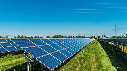 Auf einer Wiese stehen in langen Reihen Solarmodule. Im Hintergrund verläuft eine große Stromleitung.