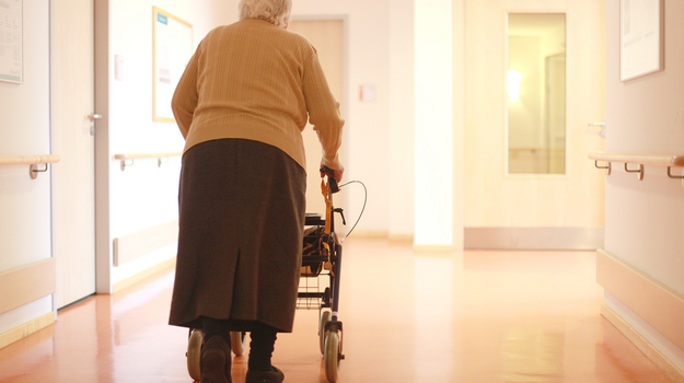 Eine ältere Frau geht mit Hilfe eines Rollators einen Gang entlang, von dem links und rechts Türen abgehen.