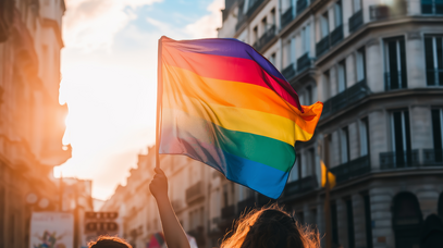Eine Person mit langen Haaren schwenkt eine Regenbogenflagge im Gegenlicht. Im Hintergrund sieht man eine Straßenzeile.