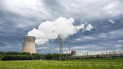 Auf der linken Bildseite stehen große Kühltürme. Auf der rechten Seite befinden sich Stromleitungen und Umspannwerke. Der Himmel ist von dunklen Wolken bedeckt.  