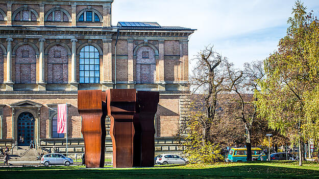 Vor einem alten Gebäude mit großen Säulen steht eine moderne Skulptur aus Metall. 