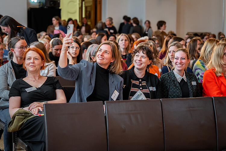 Vier Frauen sitzen auf Stühlen. Die drei rechten Frauen nehmen ein Selfie auf