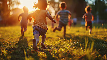 Kleine Kinder rennen auf einer Wiese dem Sonnenuntergang entgegen.