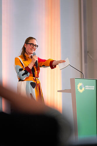 Eine Frau in buntem Kleid zeigt bei einer Rede ins Publikum