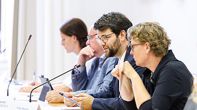 Vier Personen sitzen in einer Reihe vor Mikrofonen