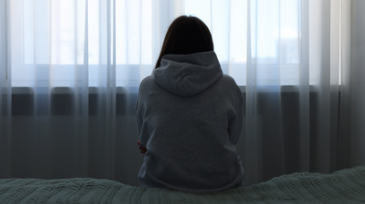 Eine Frau sitzt von der Kamera abgewandt in einem grauen Pulli auf einem Bett. Sie umschlingt ihren Körper mit ihren Armen. Das Bild wirkt düster.
