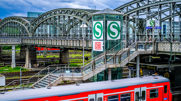 Im Vordergrund hält eine rote S-Bahn. Dahinter führt eine Treppe und ein Aufzug mit großem "S"-Symbol zu einer großen Brücke.