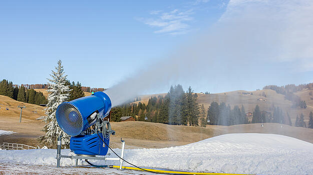 Eine Schneekanone häuft einen Schneehügel inmitten einer grünen Berglanschaft auf. 