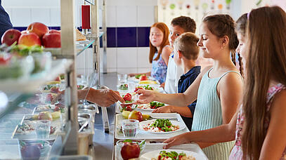 Kinder, die in der Kantine einer Schule gesunden Essen holen. Ein Mann reicht einem Mädchen eine Schale Tomaten.