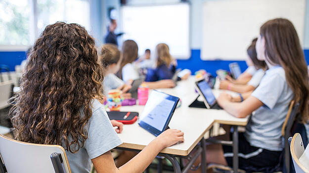 Schülerinnen sitzen in Klassenzimmer an einem Tisch zusammen vor Tablets. Vorne erklärt eine Person an der digitalen Tafel.