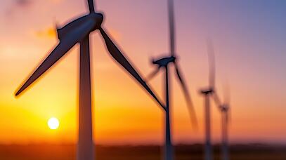 Von vorne links nach hinten rechts stehen in einer Reihe vier Windräder. Im Hintergrund geht die Sonne unter.