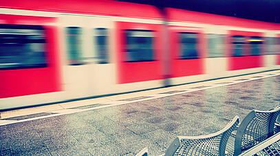 Im Hintergrund ist ein leicht verschwommener, roter Zug. Am Bahnsteig steht eine Sitzbank