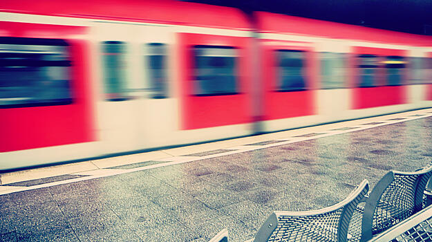 Im Hintergrund ist ein leicht verschwommener, roter Zug. Am Bahnsteig steht eine Sitzbank