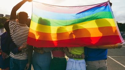 Eine Gruppe aus sechs Personen steht Arm in Arm. Ihre Köpfe sind nicht zu sehen, da sie von einer Regenbogenflagge verdeckt werden, die die Menschen gemeinsam hinter sich hochhalten