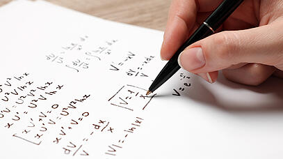 Hand schreibt mit Kugelschreiber mathematische Gleichungen auf Papier.
